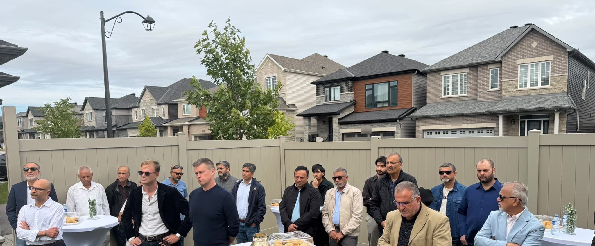 A backyard full of people standing among high tables, listening, with new houses beyond the fence