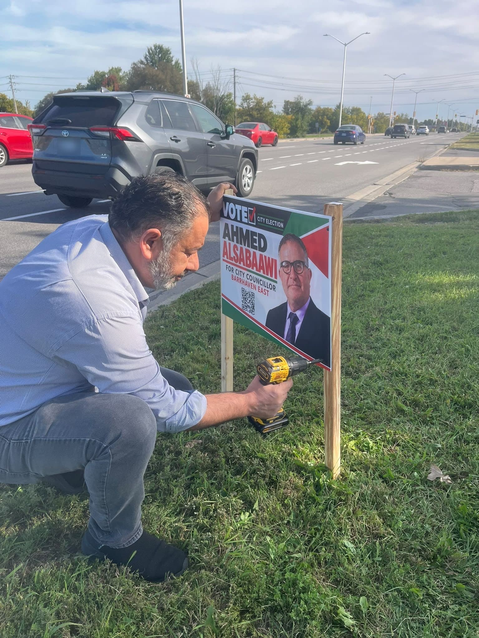 A man crouched on the grass beside a main road, fixing an Ahmed Alsabaawi lawn sign to its wooden stakes with a drill